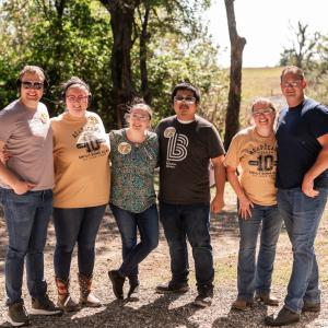 Group_of_six_people_standing_together_outdoors_with_trees_behind_them
